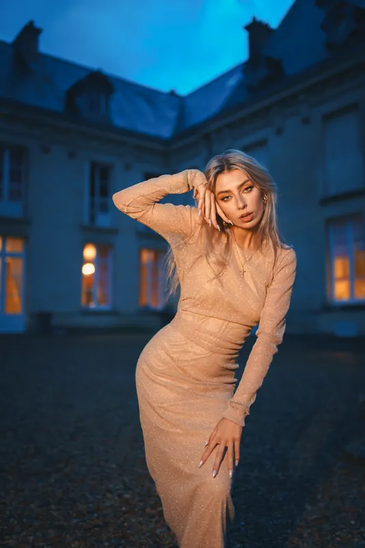 Anaïs en robe couleur sable devant la façade aux fenêtres allumées, à l’aube
